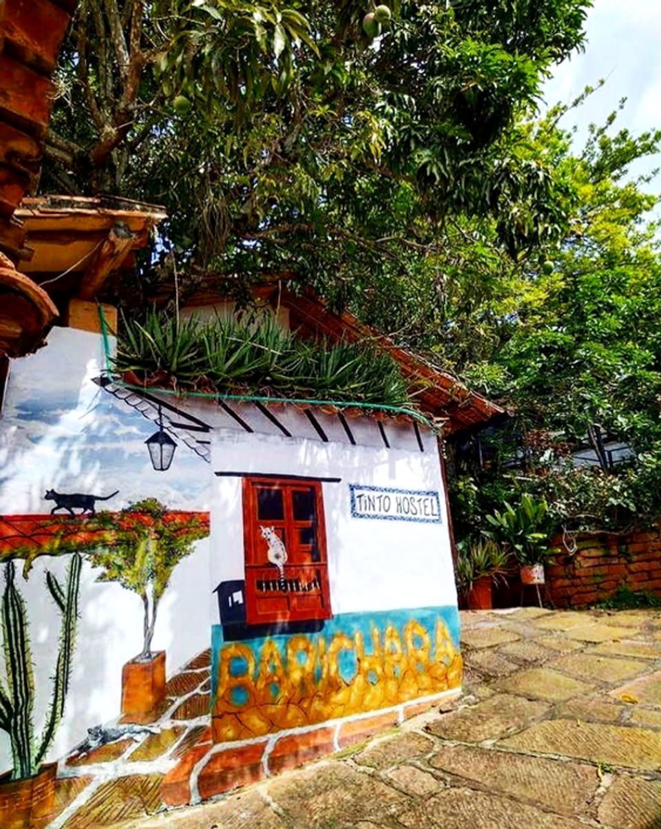 Fachada de Tinto Hostel con mural: cielo y tejado pintados con un gato negro, cactus, una ventana roja de madera, la placa de azulejo «Tinto Hostel» y la palabra «Barichara» en letras amarillas sobre una franja turquesa, junto a un camino de piedra y árboles.