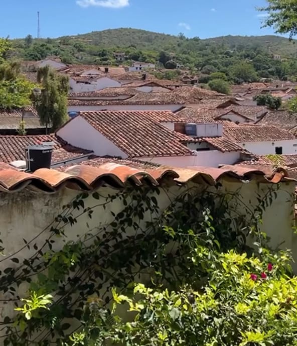 Tejados de barro de Barichara sobre un muro blanco rematado con tejas, con vegetación en primer plano y cerros verdes bajo el cielo azul.